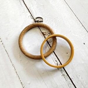 May include: Two embroidery hoops. One is made of light brown wood with a textured grain, and a metal clasp. The other is a solid, mustard-yellow plastic hoop. Both are circular and rest on a white, weathered wooden surface.