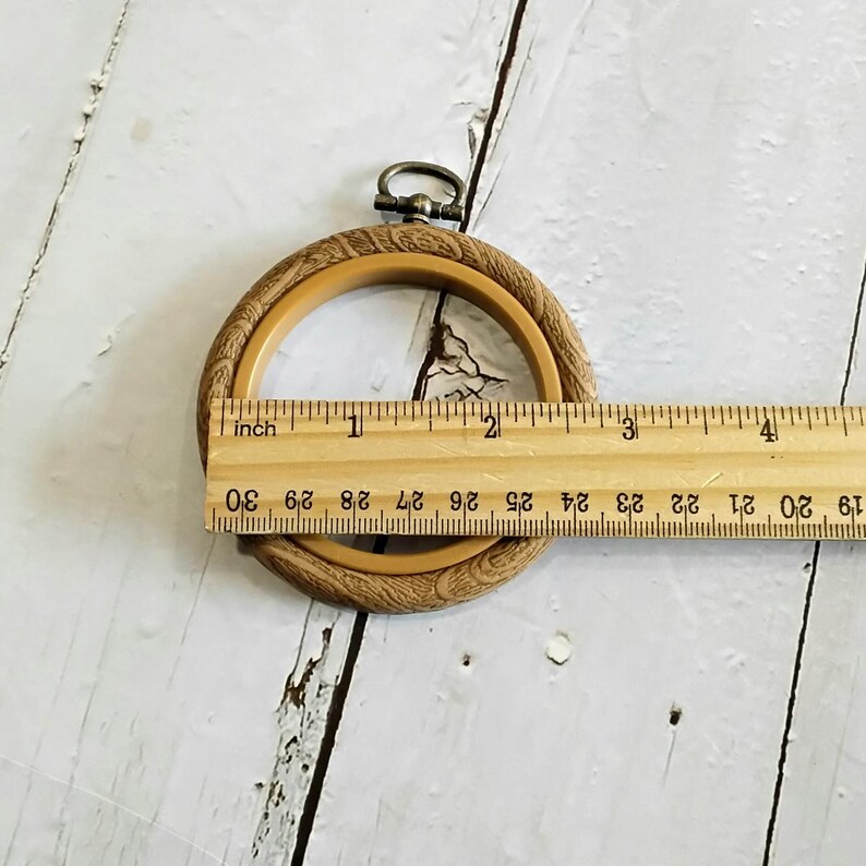 May include: A round, light brown embroidery hoop with a decorative outer edge. The hoop is approximately 10 cm in diameter. A metal clasp is at the top. A wooden ruler is placed across the hoop.