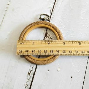 May include: A light brown embroidery hoop with a decorative outer ring. The hoop is approximately 10 cm in diameter. A metal clasp is at the top. A wooden ruler is placed across the hoop.