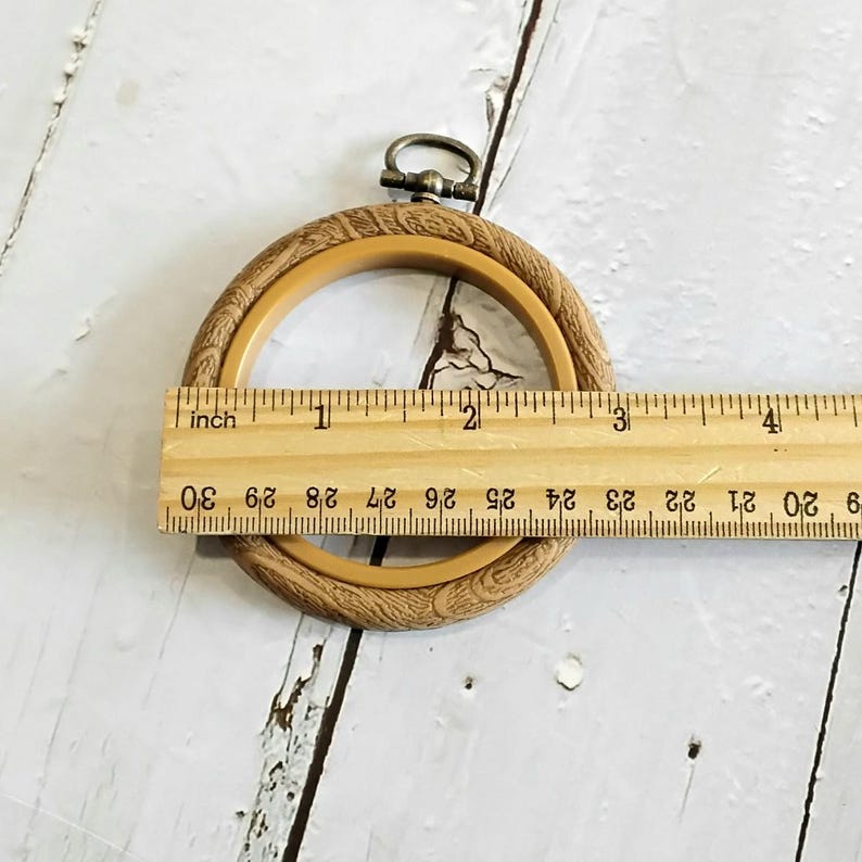 May include: A light brown embroidery hoop with a decorative outer ring. The hoop is approximately 10 cm in diameter. A metal clasp is at the top. A wooden ruler is placed across the hoop.