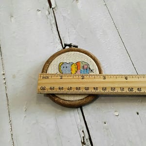 May include: A small, round cross-stitch embroidery hoop with a colourful, abstract design. The design features blue, yellow, orange, and pink thread. The wooden hoop is approximately 5 cm in diameter. A wooden ruler is placed across the hoop.