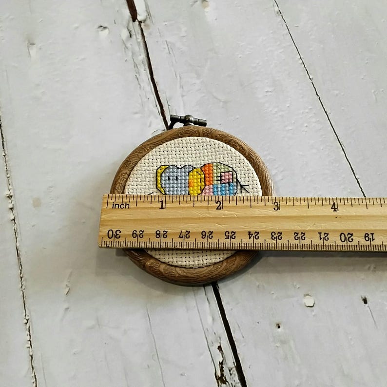 May include: A small, round cross-stitch embroidery hoop with a colourful, abstract design. The design features blue, yellow, orange, and pink thread. The wooden hoop is approximately 5 cm in diameter. A wooden ruler is placed across the hoop.