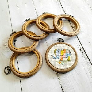 May include: A selection of wooden embroidery hoops, some empty and some with cross-stitch art. One hoop displays a colourful cross-stitched elephant design. The hoops are made of light brown wood and have a metal clasp.