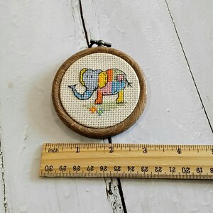 May include: A cross-stitch elephant in a wooden embroidery hoop. The elephant is stitched in blue, yellow, pink, and orange. The hoop is approximately 7.6 cm in diameter. The background is cream-colored fabric.