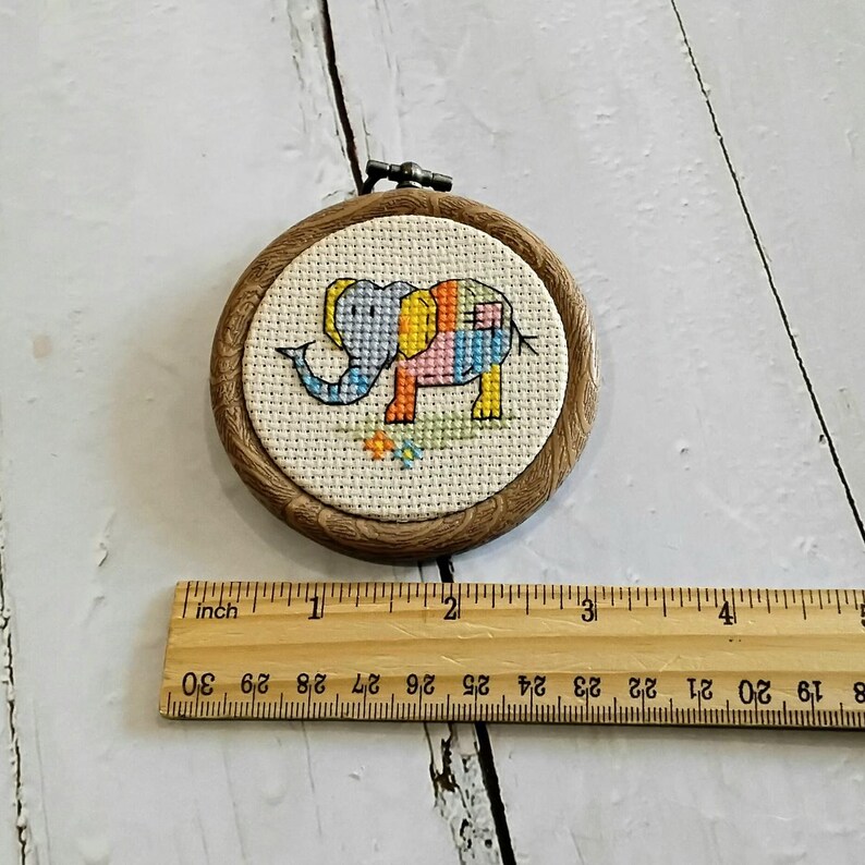 May include: A cross-stitch elephant in a wooden embroidery hoop. The elephant is stitched in blue, yellow, pink, and orange. The hoop is approximately 7.6 cm in diameter. The background is cream-colored fabric.