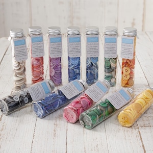 May include: A collection of twelve clear cylindrical glass tubes filled with assorted plastic craft buttons in various colours including white, red, purple, blue, green, orange, black, pink, and yellow, each labelled with a white paper tag.