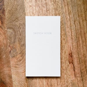 May include: A rectangular sketchbook with a plain white cover featuring the words SKETCH BOOK printed in a simple, light gray sans-serif font centered on the front. The cover has a subtle, fine-grained texture throughout its surface.