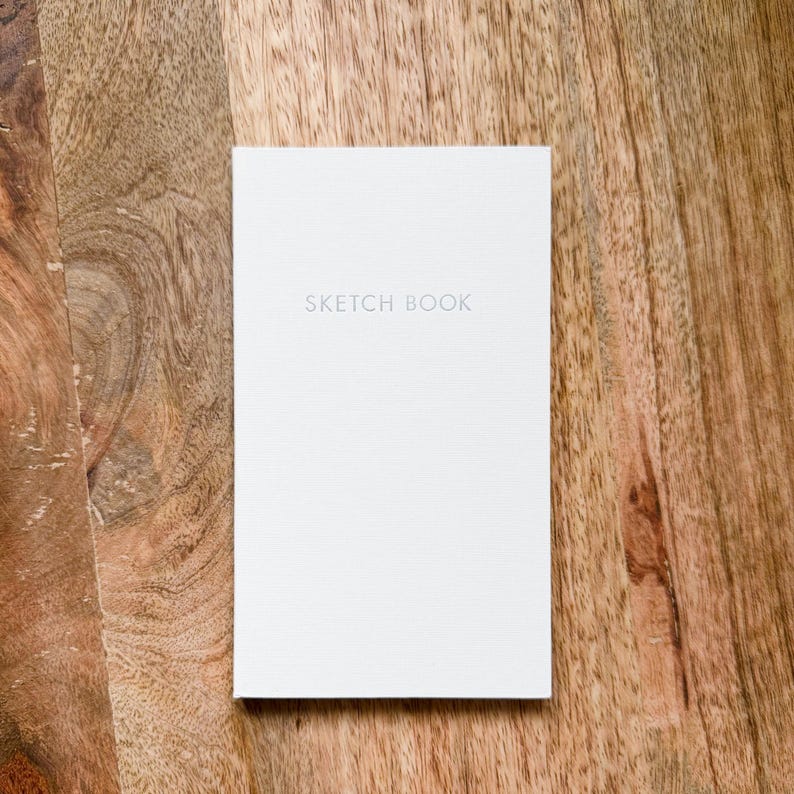 May include: A rectangular sketchbook with a plain white cover featuring the words SKETCH BOOK printed in a simple, light gray sans-serif font centered on the front. The cover has a subtle, fine-grained texture throughout its surface.