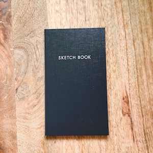 May include: A rectangular black sketchbook with a textured cover featuring the words SKETCH BOOK printed in white capital letters centered on the front. The notebook has a clean, minimalist design with a vertical orientation and a solid dark finish.