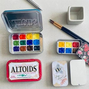 May include: A collection of watercolor paint sets in metal tins, including a large twelve-pan set, a smaller six-pan set with a floral lid, a red Altoids peppermint tin, a plain metal tin, two paintbrushes, a small ceramic water cup, and a pen.