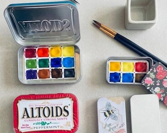 May include: A collection of watercolor paint sets in metal tins, including a large twelve-pan set, a smaller six-pan set with a floral lid, a red Altoids peppermint tin, a plain metal tin, two paintbrushes, a small ceramic water cup, and a pen.