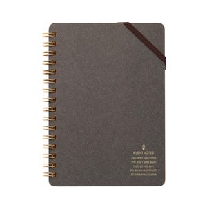 May include: A dark gray spiral-bound notebook with gold-colored metal rings. The notebook has a brown elastic band closure and the text "KLEID NOTES" is visible. The notebook is A6 size and contains 70 sheets.