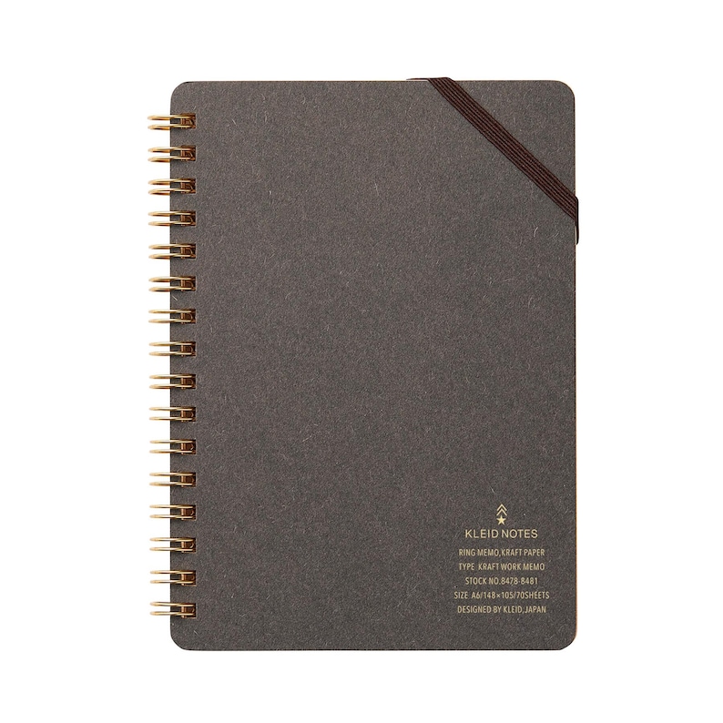 May include: A dark gray spiral-bound notebook with gold-colored metal rings. The notebook has a brown elastic band closure and the text "KLEID NOTES" is visible. The notebook is A6 size and contains 70 sheets.
