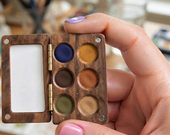 May include: A small, rectangular, hinged wooden watercolor palette featuring a dark wood grain finish. The palette contains six circular wells filled with solid paint in shades of navy blue, mustard yellow, dark brown, terracotta, olive green, and tan.