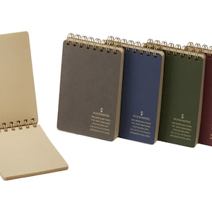 May include: A collection of spiral-bound notebooks in various colors, including gray, blue, green, and burgundy. One notebook is open, revealing grid-lined pages. The notebooks have gold-colored spiral bindings and text on the covers.