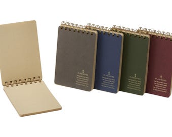 May include: A collection of spiral-bound notebooks in various colors, including gray, blue, green, and burgundy. One notebook is open, revealing grid-lined pages. The notebooks have gold-colored spiral bindings and text on the covers.