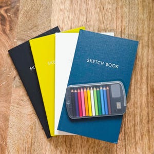 May include: Four rectangular sketchbooks in black, yellow, white, and blue with the text SKETCH BOOK printed on the covers. A clear plastic case containing ten colored pencils and a small eraser is placed on top of the blue sketchbook.