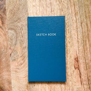 May include: A rectangular sketchbook with a textured deep blue cover. The words SKETCH BOOK are printed in white, capitalized, sans-serif letters across the upper center of the front cover. The notebook has a clean, vertical orientation.