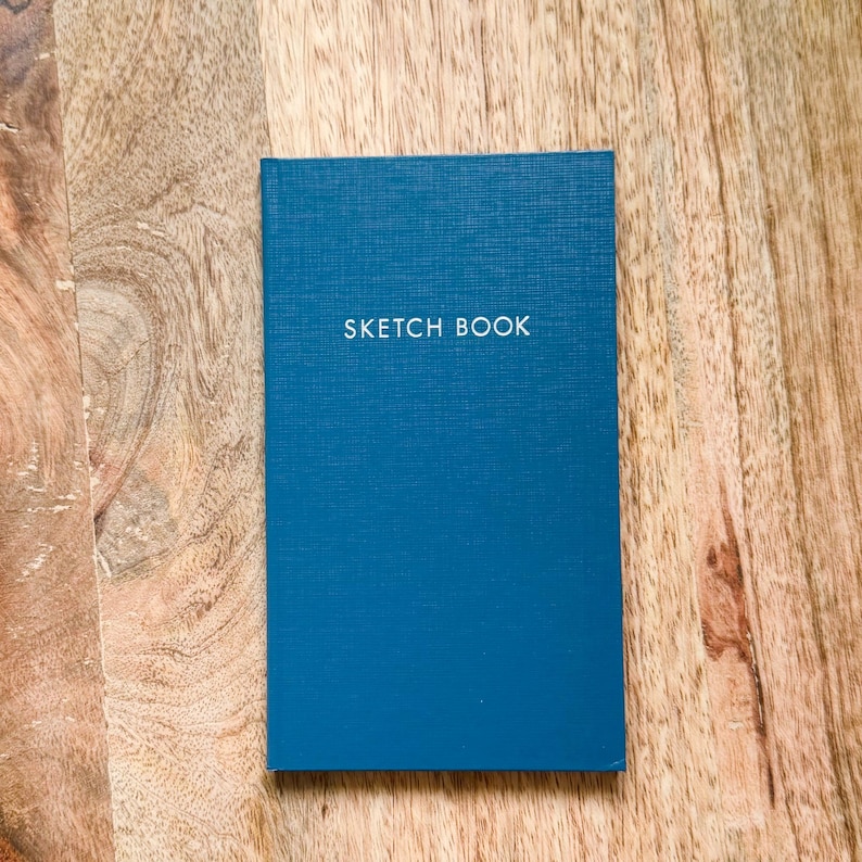 May include: A rectangular sketchbook with a textured deep blue cover. The words SKETCH BOOK are printed in white, capitalized, sans-serif letters across the upper center of the front cover. The notebook has a clean, vertical orientation.