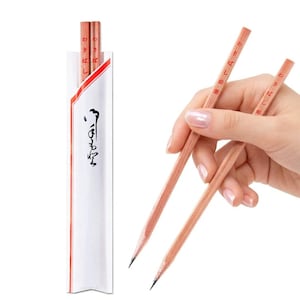 May include: Wooden chopsticks with pencil tips, held in a hand. The chopsticks are light brown with Japanese text. A white paper sleeve with red trim and Japanese calligraphy contains another set of chopsticks.