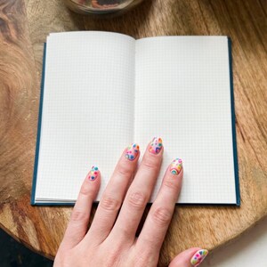 May include: An open notebook with white grid paper pages and a dark blue cover. The pages are blank and feature a light gray grid pattern. The notebook is positioned flat, displaying two open pages ready for writing or drawing.