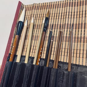 May include: A collection of calligraphy brushes in a black fabric roll, displayed on a bamboo mat. The brushes vary in size and shape, with wooden handles and different colored bristles. The roll has a red border.