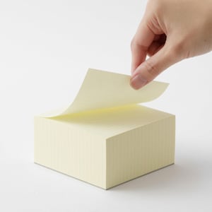 May include: A square stack of pale yellow adhesive notes with a subtle grid pattern. The top sheet is partially lifted, revealing the light-colored paper. The stack is thick and maintains a uniform, block-like shape with clean, straight edges.