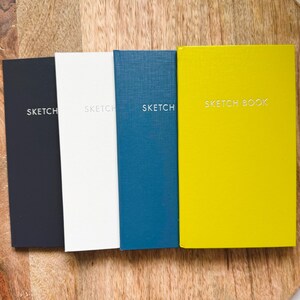May include: A set of four rectangular sketchbooks in black, white, blue, and yellow. Each cover features a textured finish and the word SKETCH or SKETCH BOOK embossed in white lettering on the front. The books are arranged side by side.