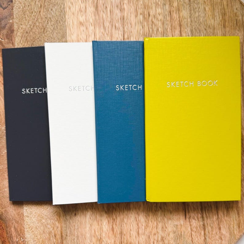 May include: A set of four rectangular sketchbooks in black, white, blue, and yellow. Each cover features a textured finish and the word SKETCH or SKETCH BOOK embossed in white lettering on the front. The books are arranged side by side.