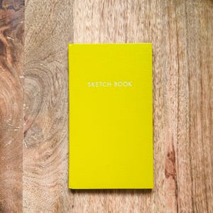 May include: A rectangular sketchbook with a vibrant lime green cover featuring the words SKETCH BOOK printed in white capital letters centered on the front. The notebook has a clean, solid-colored finish and a simple, functional design.