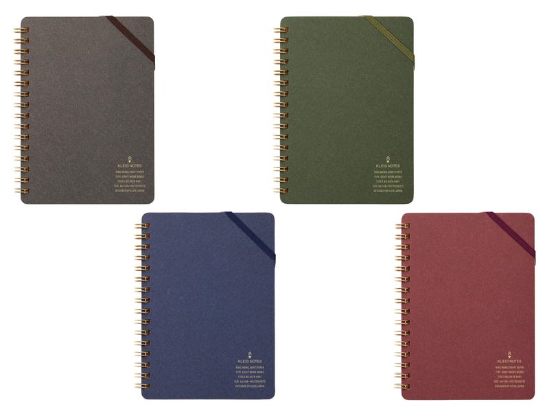 May include: A collection of spiral-bound notebooks in various colors: charcoal, olive green, navy blue, and burgundy. Each notebook features a gold spiral binding, a matching elastic closure, and a small printed logo with text.