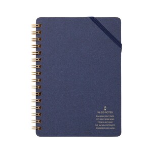 May include: A navy blue spiral-bound notebook with gold-colored metal rings. The notebook has an elastic band closure and the text "KLEID NOTES" is printed on the bottom right corner. The notebook is designed in Japan.