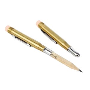 May include: Two brass-colored metal bullet-shaped pencil holders with silver-toned clips and pink erasers. One holder contains a wooden pencil with a visible signature, while the other is capped with a silver-toned tip. The design mimics a cartridge.
