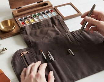 May include: A dark brown canvas roll-up case with multiple slots containing paintbrushes and a pen. Beside it sits a wooden watercolor palette box with various paint pans, a small brass bowl, and a square wooden frame on a white surface.