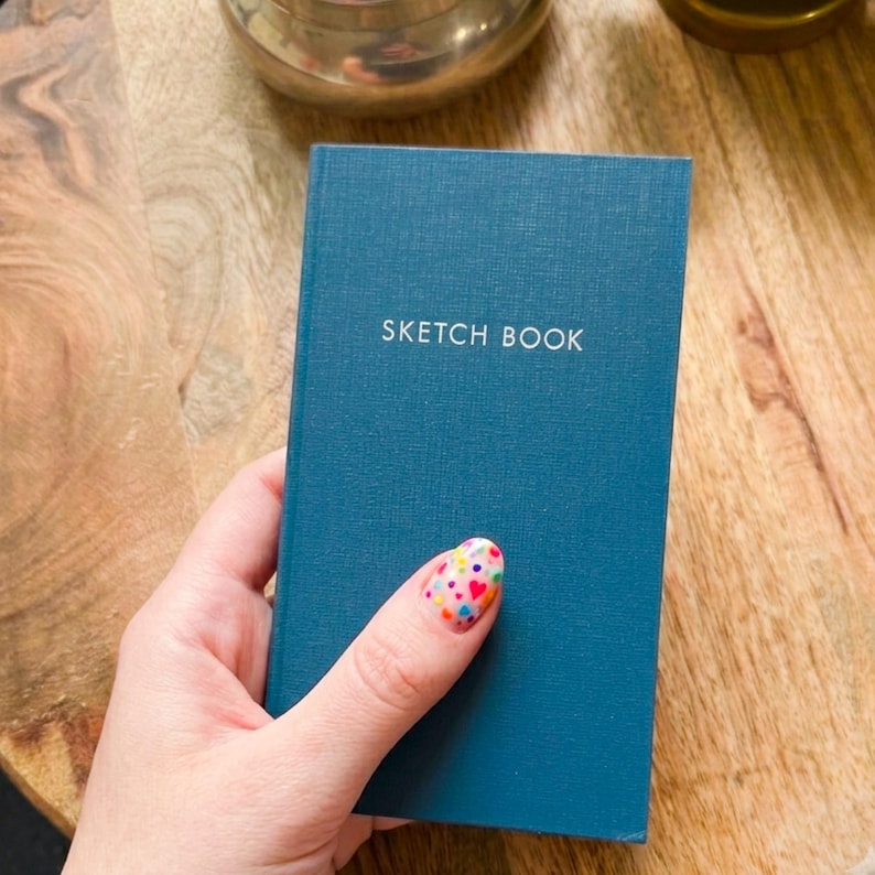 May include: A rectangular sketchbook with a solid dark teal fabric cover and a textured surface. The words SKETCH BOOK are printed in white capital letters on the front cover. The book has a vertical orientation and a clean, simple design.