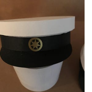 May include: A white and black graduation cap with a gold emblem. The cap features a circular gold emblem with a star design. The top of the cap is white, with a black band in the middle.