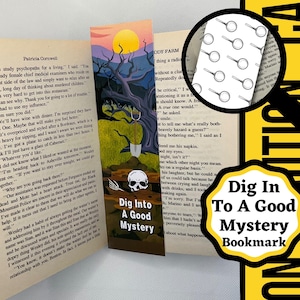 May include: A colorful bookmark with the text "Dig Into A Good Mystery." The bookmark features a spooky illustration of a skull, a shovel, and a tree under a yellow sun. The bookmark is placed in an open book.