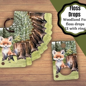 以下が含まれることがあります： リング付きの「Woodland Fox」刺繍糸プレゼンテーション。デザインは、キツネ、切り株、森の風景の水彩イラストが特徴です。「Floss Drops」と「Woodland Fox floss drops 12 with ring」のテキストが表示されています。