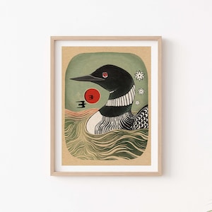 May include: A framed art print featuring a black loon with a red eye swimming in green water. The illustration includes a red sun, a small black bird silhouette, and three white star-like shapes against a textured tan background.