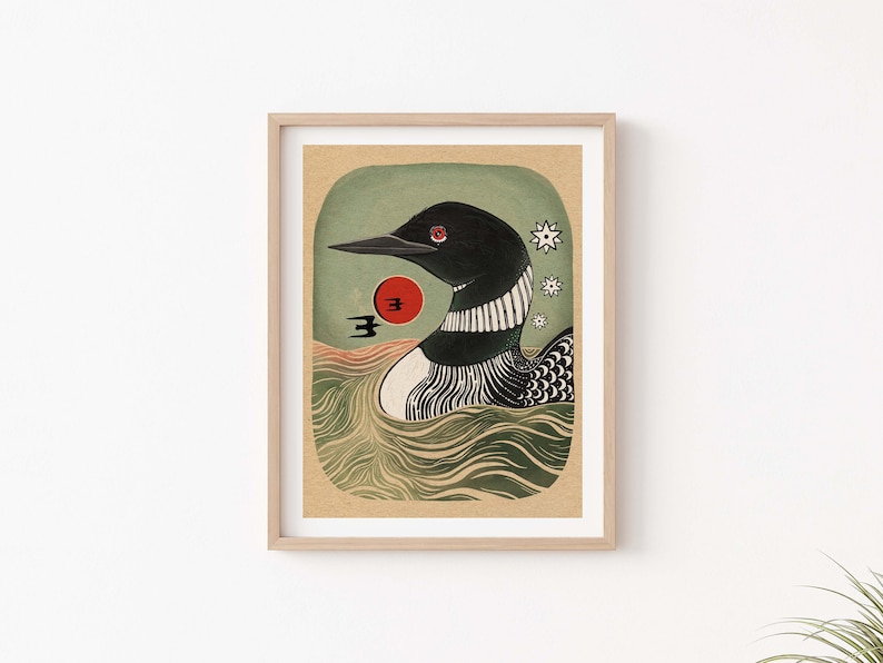 May include: A framed art print featuring a black loon with a red eye swimming in green water. The illustration includes a red sun, a small black bird silhouette, and three white star-like shapes against a textured tan background.