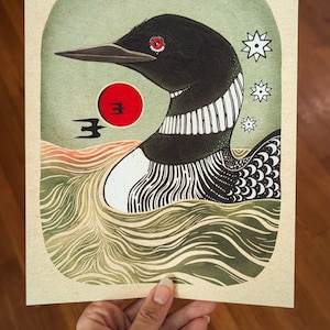 May include: A rectangular art print featuring a black loon with a red eye swimming in green water. A bright red sun sits behind the bird, accompanied by a small black bird silhouette and three white star shapes on a light cream background.