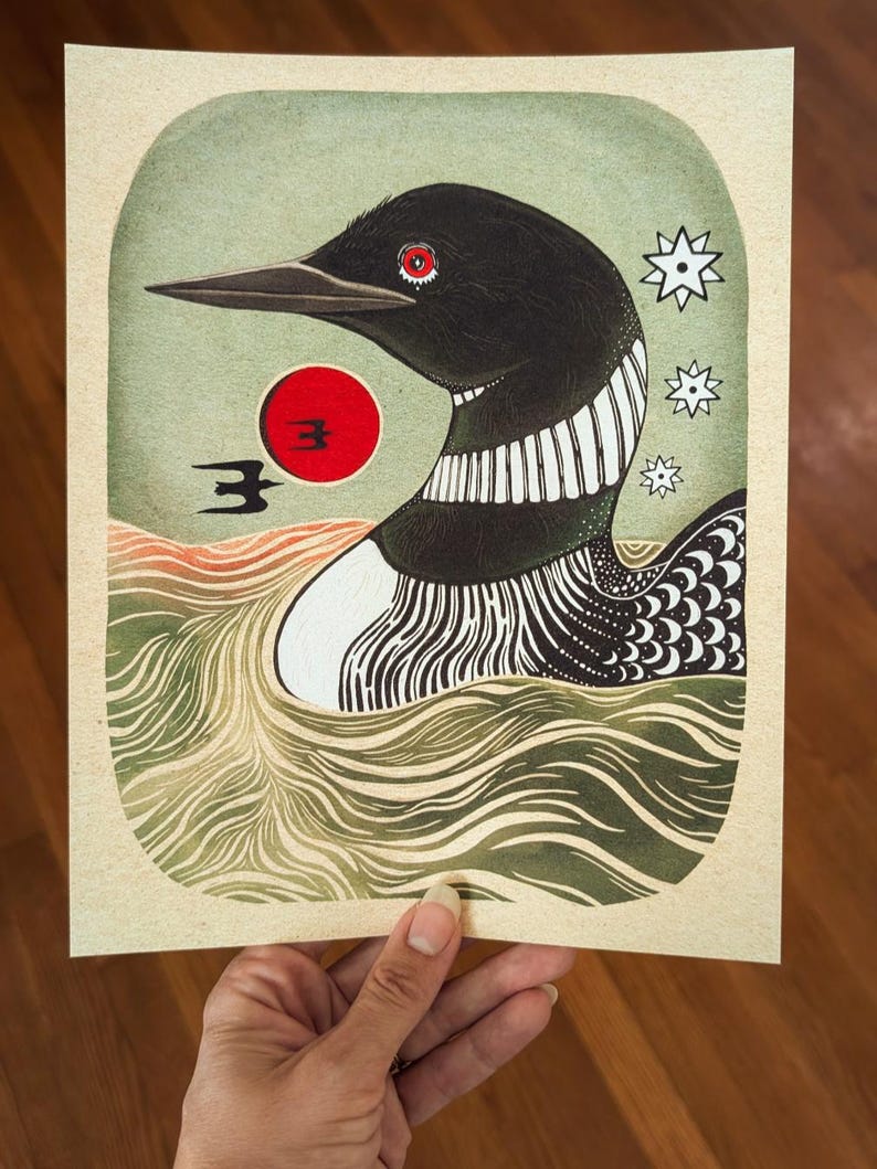 May include: A rectangular art print featuring a black loon with a red eye swimming in green water. A bright red sun sits behind the bird, accompanied by a small black bird silhouette and three white star shapes on a light cream background.