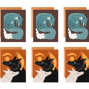 May include: A set of six greeting cards featuring two distinct designs. The top row shows a blue fox curled in a brown landscape with a crescent moon. The bottom row displays a black fox and a white fox against an orange background with a comet.