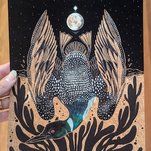 May include: A rectangular art print featuring a black and metallic gold illustration of a loon with wings spread wide. The bird has a white-spotted back, a dark head with a red eye, and is positioned below a moon and a star against a black sky.