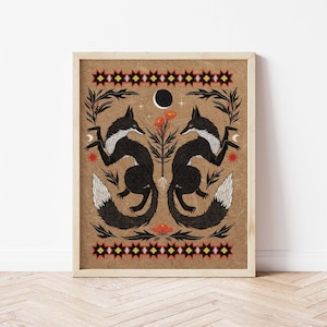 May include: Framed art print featuring two stylized black foxes facing each other, with white accents and tails. The artwork includes floral and celestial elements, set against a tan background with decorative borders. The print is in a wooden frame.