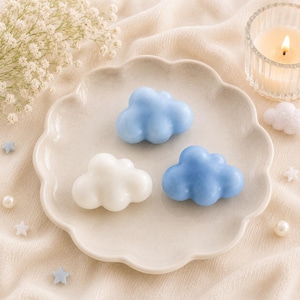May include: Three cloud-shaped soaps, two in light blue and one in white, are arranged on a scalloped-edge plate. A lit candle in a glass holder and decorative stars and pearls are in the background. The soaps are likely for a baby or nursery theme.