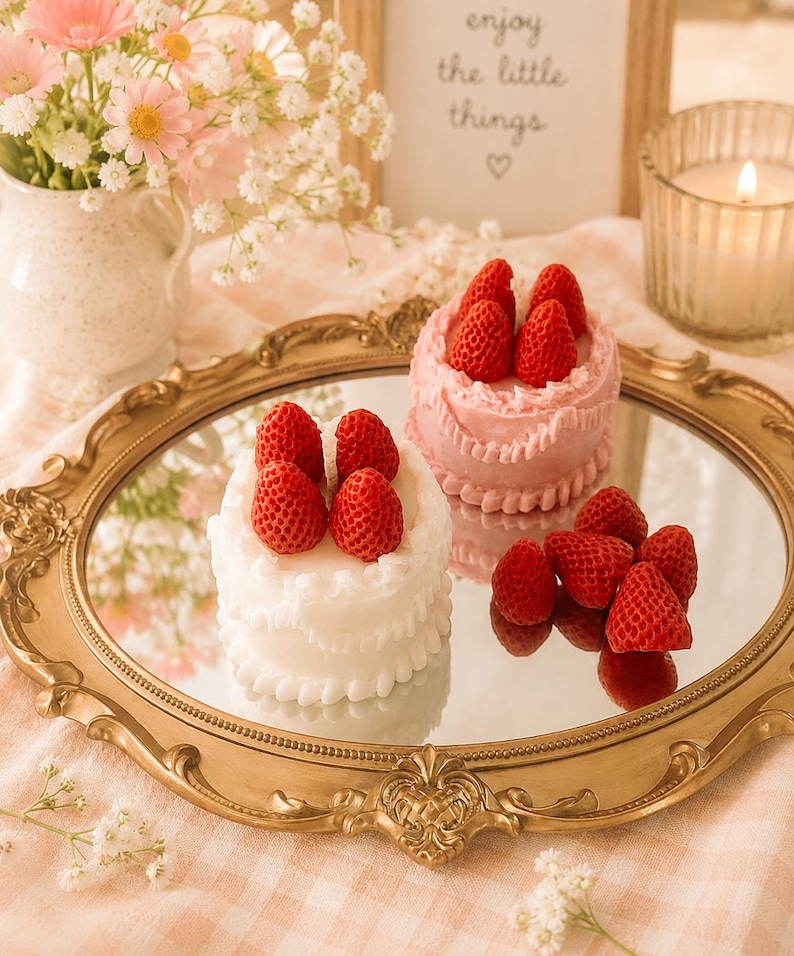 May include: A gold-framed mirror holds two small, round cakes topped with fresh strawberries. One cake is white, the other pink. The mirror sits on a pink and white checkered cloth, with flowers and a candle in the background.