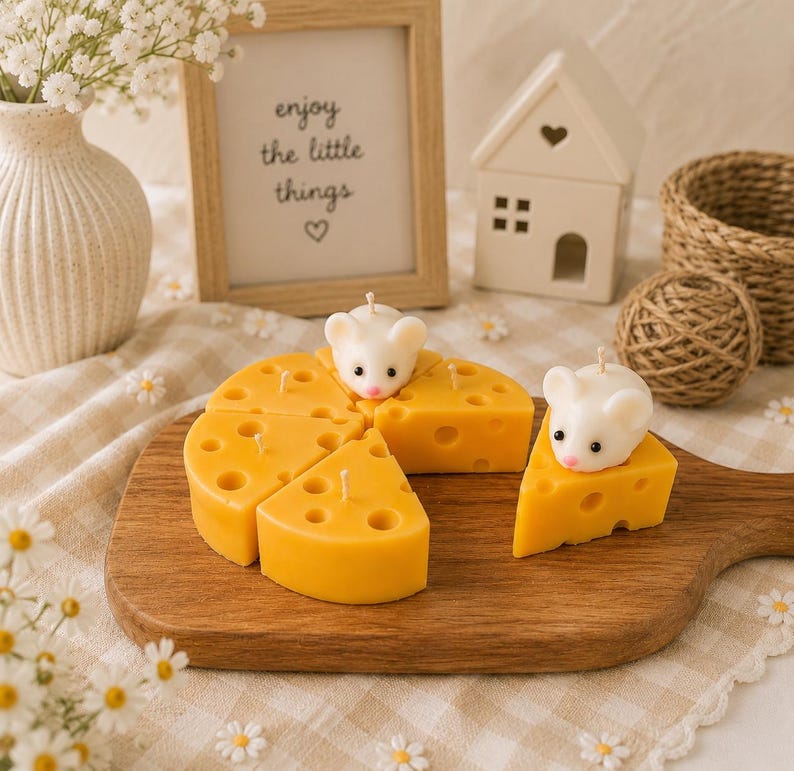 May include: A wooden serving board displays a cheese-shaped candle cut into wedges, each with holes. Two white mouse candles sit atop the cheese. A framed sign reads "enjoy the little things."