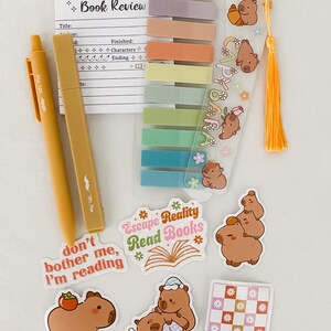May include: A collection of stationery items, including a yellow pen, sticky notes, stickers, and a bookmark with a tassel. The stickers feature capybaras and phrases like "Escape Reality Read Books." The bookmark has the text "Happy Capybara".