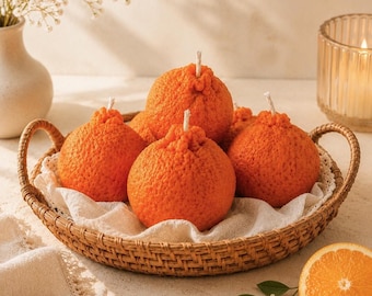 Puede incluir: Una cesta de mimbre contiene cinco velas de color naranja con forma de naranjas, cada una con una mecha blanca. La cesta descansa sobre un paño blanco. Un jarrón con flores blancas y una vela encendida están en el fondo. También se ve una naranja en rodajas.